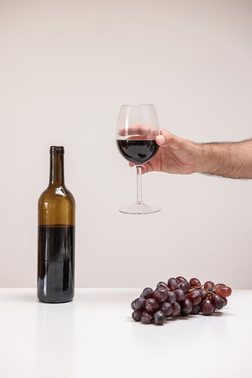 A hand holding a glass of red wine next to a bottle and fresh grapes on a white table.